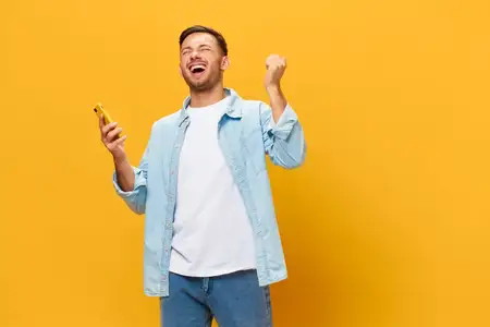Необычайно счастливый загорелый красивый мужчина в синей базовой футболке поднимает кулак вверх, используя телефон, позирует изолированно на оранжево-желтом студийном фоне