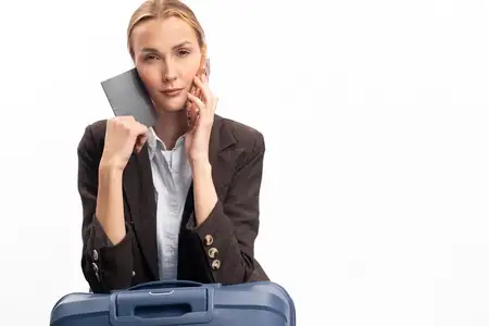 Уверенная молодая женщина в коричневом пиджаке с паспортом и смартфоном кладет руки на синий чемодан