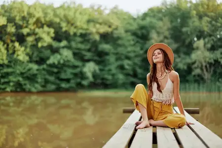 Женщина-путешественница, хиппи и экоактивистка, сидит на мосту у озера, раскинув руки, в шляпе и искренне улыбается
