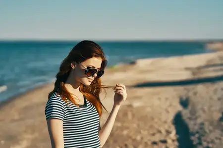Женщина в футболке и солнцезащитных очках стоит на песчаном берегу пляжа у моря