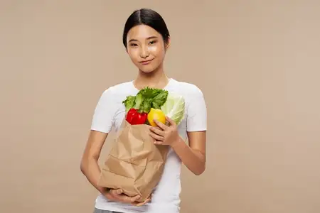 Стройная женщина с пакетом продуктов в руке на бежевом фоне, обрезанный вид, диета
