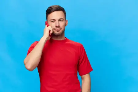 Счастливый задумчивый загорелый симпатичный мужчина в красной футболке разговаривает с семьёй по телефону, смотрит в сторону и позирует изолированно на синем студийном фоне