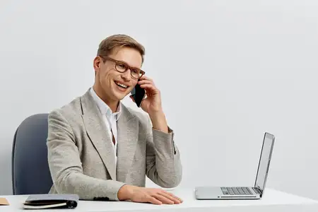 Улыбающийся молодой мужчина европеоидной расы разговаривает по смартфону, сидя за столом с ноутбуком в современном минималистичном офисе