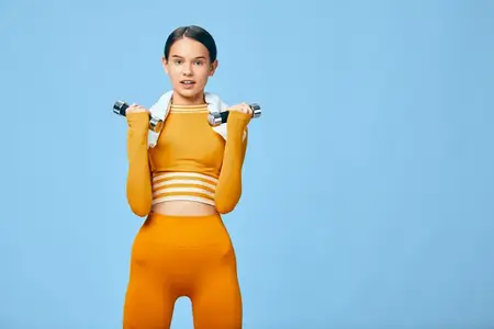 Спортсменка тренируется в современной спортивной одежде, выполняя сгибание рук с легкой гантелью, в топе-кропе и леггинсах