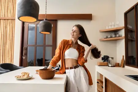 Улыбающаяся молодая женщина в уютной современной кухне, демонстрирующая здоровый образ жизни, одетая в стильный наряд, играет со своими волосами при естественном освещении