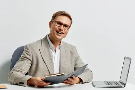 Жизнерадостный мужчина в повседневной деловой одежде работает за столом с ноутбуком и документами в светлом современном офисе, демонстрируя уверенность и профессионализм, наслаждаясь своей работой