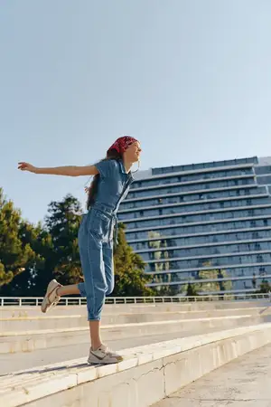 Женщина в стиле life style, одетая в современный бохо-вестерн деним стритстайл, наслаждается балансированием на ступенях с раскинутыми руками в окружении городской архитектуры и зелени в теплом…