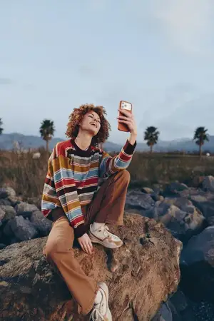 Женщина на открытом воздухе делает селфи на смартфон, на ней вьющиеся волосы и полосатый свитер