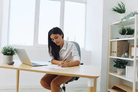 Женщина работает и учится, записывая текст на бумаге ручкой и проверяя его на ошибки, занимаясь учебой и преподаванием в колледже, выполняя бизнес-работу за столом с ноутбуком