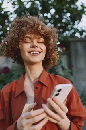 Улыбающаяся женщина на улице пользуется смартфоном, одетая в повседневную одежду в солнечный день
