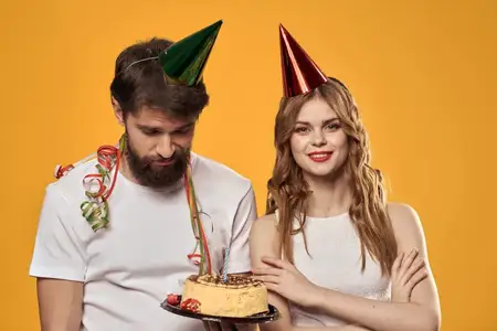 Счастливые мужчина и женщина в колпаках празднуют день рождения на жёлтом фоне, держа в руках торт