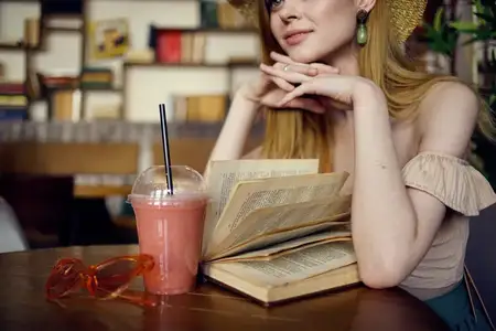 Женщина в шляпе сидит в кафе, перед ней стакан с напитком и книга для отдыха