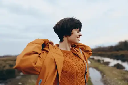Женщина на отдыхе на природе: в оранжевой куртке идет вдоль береговой линии, на заднем плане вид на океан, улыбается на ветру, атмосфера путешествия на природе, открытое небо и далекие холмы