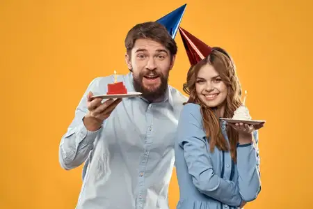 Мужчина и женщина на праздновании дня рождения на жёлтом фоне, в колпаках, с тортом в руках
