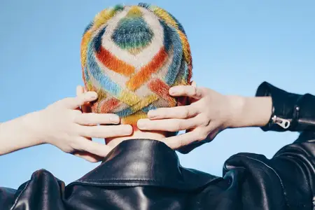 Женщина, одетая в черную кожаную куртку и яркую разноцветную вязаную шапку, стоит, положив руки на голову, на фоне яркого голубого неба