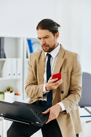 Мужчина в бежевой куртке держит телефон и ноутбук в офисе, используя технологии