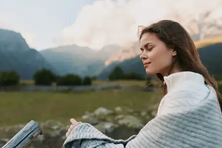 Безмятежная, задумчивая женщина наслаждается свежим воздухом в фактурном свитере, с дальними горами и голубым небом, создающими естественную лайфстайл-сцену