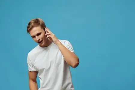 Улыбающийся молодой мужчина европеоидной расы разговаривает по смартфону, одетый в повседневную белую футболку, изолирован на синем фоне со счастливым выражением лица