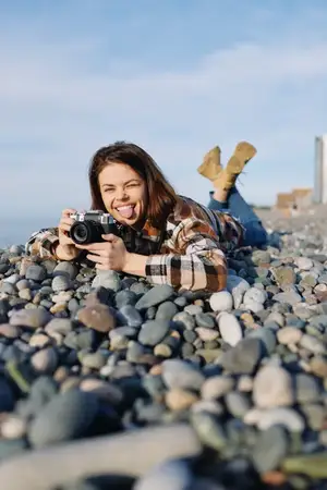 Непринужденный снимок улыбающейся женщины-фотографа на галечном пляже: она снимает пейзаж камерой, расслабленный стиль одежды и прибрежная атмосфера при дневном свете