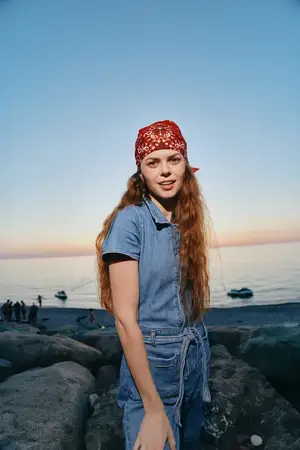 Молодая женщина в красной бандане и джинсовой одежде стоит на каменистом пляже во время спокойного заката у моря, с мягким естественным освещением и расслабленным настроением