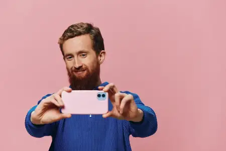 Портрет рыжеволосого мужчины с телефоном в руке, который делает селфи и фотографирует на свой телефон с улыбкой на розовом фоне