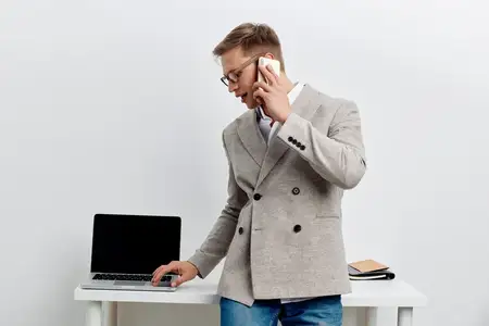 Молодой мужчина европеоидной расы работает за ноутбуком и смартфоном, разговаривая по телефону, стоит возле стола в современном офисе в повседневной куртке и очках
