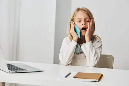 Улыбающаяся девушка-подросток занимается дома за ноутбуком в наушниках, окружённая книгами и тетрадями