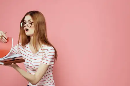 Молодая студентка с книгами на розовом фоне, в очках, крупный план лица