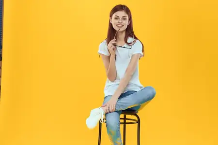 Женщина сидит на стуле и держит в руках темные очки, на ней белая футболка, женская модная одежда, желтый фон