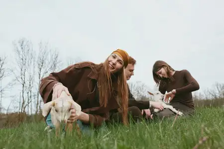 Молодая женщина в коричневой куртке и оранжевой повязке на голове держит козлёнка на зелёном поле, в то время как мужчина и женщина взаимодействуют с другим козлом, создавая расслабленную жизненную…