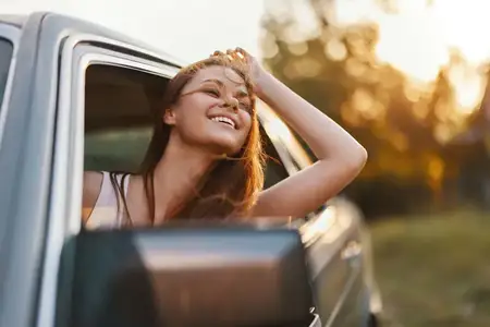 Веселая молодая женщина выглядывает из окна автомобиля, улыбаясь и наслаждаясь закатом