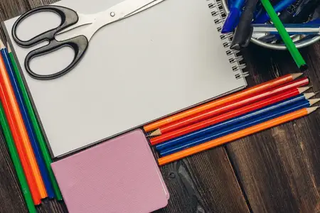 Блокноты, цветные карандаши и канцелярские принадлежности в стакане на деревянном столе, вид сверху