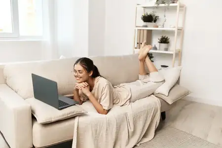 Современная женщина улыбается и работает за ноутбуком в помещении, в уютной гостиной