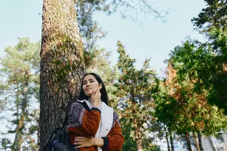 Женщина стоит рядом с деревом среди лесного света, в трикотаже и повседневной одежде, образ жизни на открытом воздухе, связь с природой, спокойная и безмятежная сцена на свежем воздухе