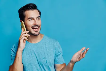 Веселый улыбающийся мужчина ведет оживленный разговор по мобильному телефону, одетый в повседневную голубую футболку, излучая положительную энергию на фоне ярко-голубого фона