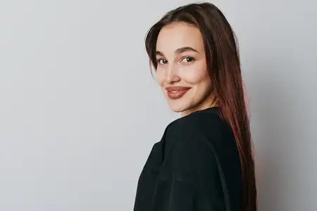 Портрет улыбающейся женщины с прямыми волосами в черном топе, оглядывающейся на камеру