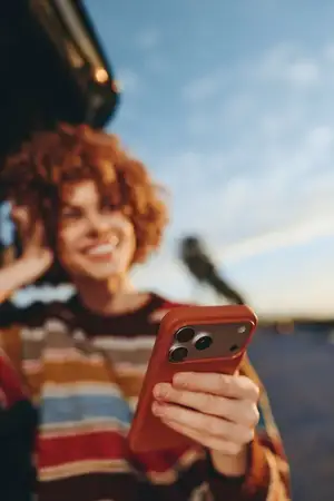 Женщина на открытом воздухе, вьющиеся волосы, держит смартфон, смеется с яркой улыбкой, залитый солнцем теплый час света, повседневный полосатый свитер, расслабленная поза, мягкий размытый фон…
