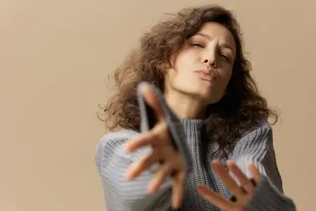 Дружелюбная кокетливая кудрявая красивая девушка в сером теплом повседневном свитере тянет руки и посылает воздушный поцелуй в камеру, позируя изолированно на пастельно-бежевом фоне