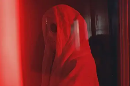 Таинственная фигура в балахоне с капюшоном отражается в окне с красным светом на темном фоне ночью