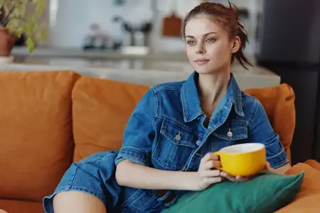 Женщина расслабляется на диване, держа в руке чашку кофе, и наслаждается уютным моментом в джинсовой рубашке