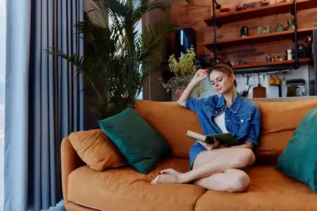 Расслабленная женщина сидит на диване с книгой, а перед ней стоит растение в уютной гостиной