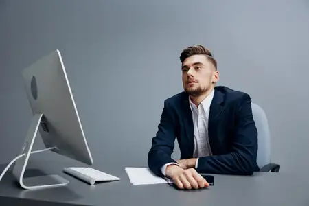 Красивый бизнесмен с проблемами на работе сидит за столом перед компьютером на сером фоне
