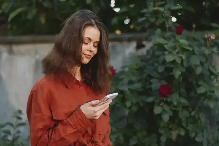 Повседневный портрет женщины на улице: смотрит в телефон, теплые цвета, цветущие розы, зелень на заднем плане, спокойное выражение лица, повседневный момент, аутентичный образ жизни