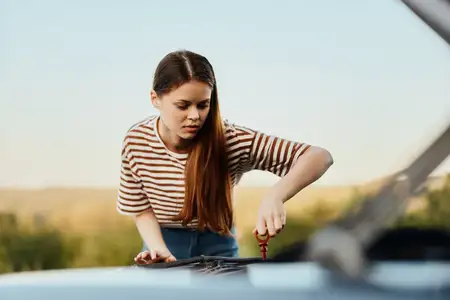 Женщина-водитель, у которой сломалась машина, вытаскивает щуп и проверяет уровень моторного масла на дороге в одиночестве