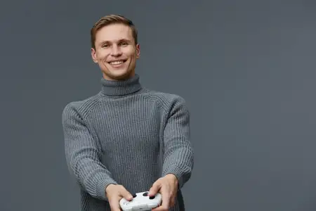 Улыбающийся мужчина держит игровой контроллер, одетый в серую водолазку, изолирован на сером фоне