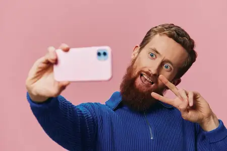 Портрет рыжеволосого мужчины с телефоном в руке, который делает селфи и фотографирует на свой телефон с улыбкой на розовом фоне