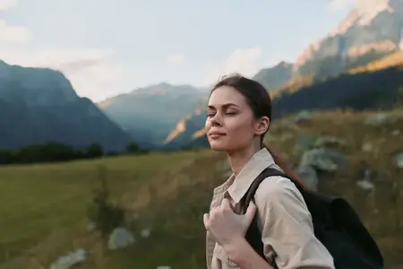 Молодая женщина в бежевой рубашке и темном рюкзаке стоит на живописном альпийском лугу, излучая спокойную уверенность и готовность к приключениям при ясном дневном свете, ее поза предполагает баланс…