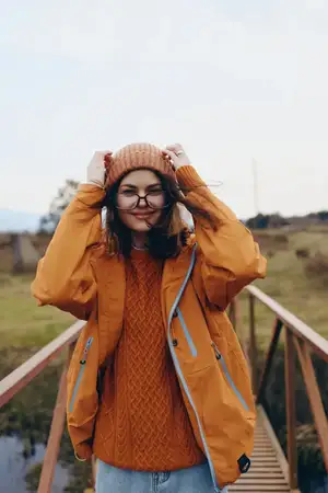 Женщина на отдыхе на природе осенью: оранжевое пальто, вязаная шапка, очки, свитер, идет по мосту в сельской местности, повседневные теплые слои одежды, руки подняты, поправляет шапку, расслабленная…