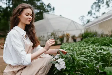 Женщина наслаждается временем в теплице, одетая в повседневную светлую одежду, в окружении ярких цветов, что представляет темы садоводства, радости и признательности природе