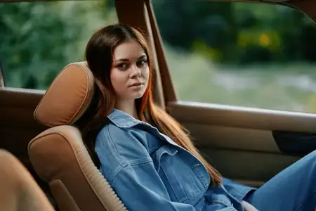 Безмятежная молодая женщина в джинсовой куртке и джинсах отдыхает в машине в окружении зелени, демонстрируя спокойный образ жизни и современную эстетику моды